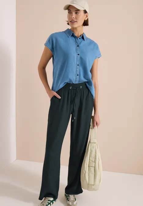 High Waist straight legs broek in Loose Fit palm breeze green Vrouw in jeansblouse, zwarte wijdvallende broek, witte pet en beige tas tegen neutrale achtergrond.
