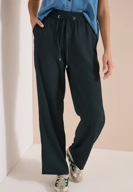 High Waist straight legs broek in Loose Fit palm breeze green Zwarte, wijde broek met trekkoord in de taille, gecombineerd met witte sneakers en een blauw shirt.