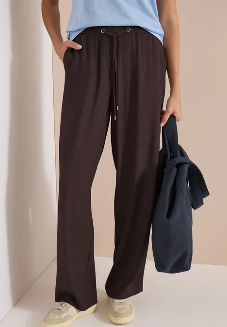 Vrouw draagt wijde donkerbruine broek, lichtblauw shirt, witte sneakers, met een donkerblauwe tas in haar hand.