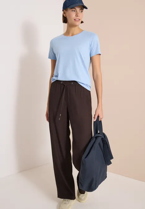 Een persoon met lichtblauw t-shirt en bruine broek, draagt een donkerblauwe tas.