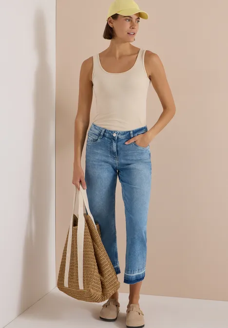 Straight Leg Jeans mit frasigem Saum light blue washed Frau mit weißem Top, hochgekrempelten Jeans, beiger Cap, Sneakers und passender Tasche vor schlichtem Hintergrund.