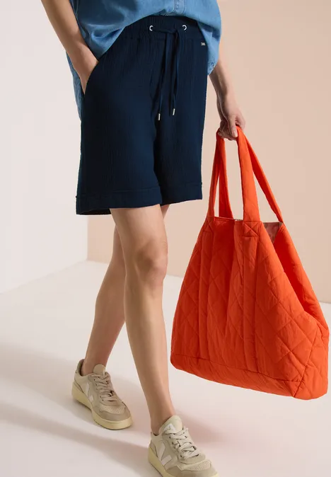 Beinpartie in dunkelblauen Shorts hält großen orangefarbenen Steppbeutel, trägt Sneaker.