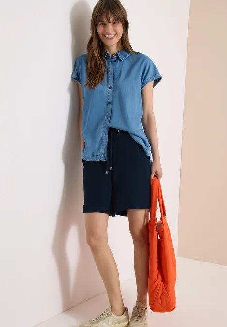 Frau lehnt an Wand und trägt blaues Hemd, dunkle Shorts und orangefarbene Tasche.