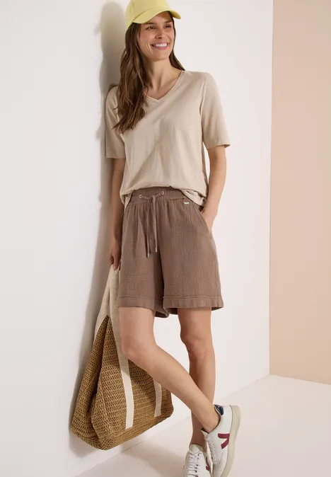 Vrouw leunend tegen muur met beige top, bruine short, witte sneakers en gevlochten tas.