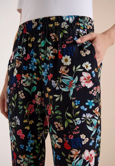 Schwarze, locker sitzende Hose mit buntem Blumenmuster, Hand in seitlicher Tasche.