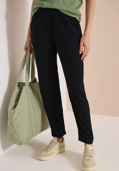 Persoon draagt donkerblauwe broek, groen shirt, heeft een doorgestikte groene shopper en beige sneakers.