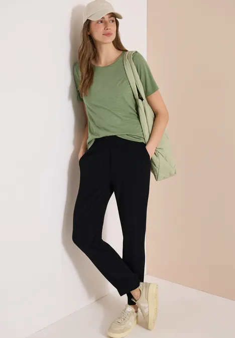 Een vrouw leunt tegen een muur, met een groen t-shirt, zwarte broek, witte schoenen en een groene tas.