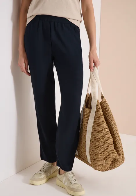 Person trägt dunkelblaue Hose, beigefarbenes Oberteil, Sneakers und hält große geflochtene Strohtasche.