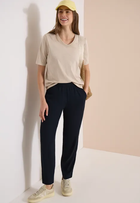 Frau lehnt entspannt an Wand, trägt beiges T-Shirt, dunkelblaue Hose und beige Sneaker.