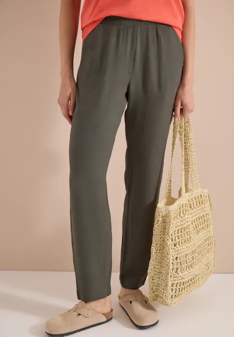 Person trägt olivgrüne Jogginghose, beige Slipper und hält eine geflochtene Strohtasche.