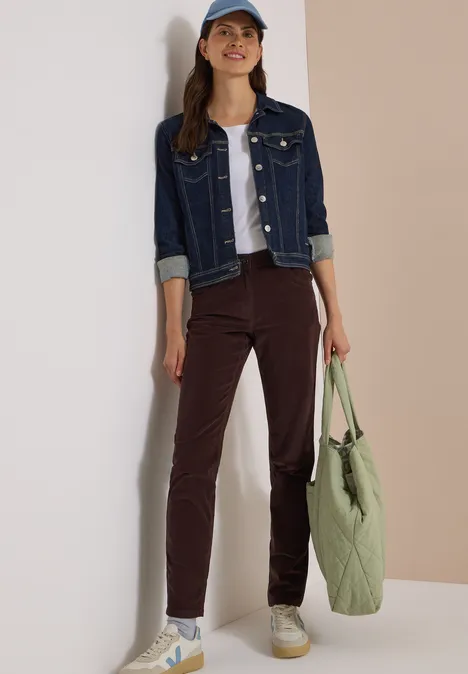 Eine Frau in Jeansjacke, weißem Top, brauner Hose und Cap lehnt mit grünem Beutel an einer Wand.