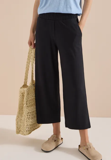 3/4 Wide Leg Hose im Casual Fit Black Schwarze, weit geschnittene Culotte mit hohem Bund, kombiniert mit Basttasche und beigen Slippern.