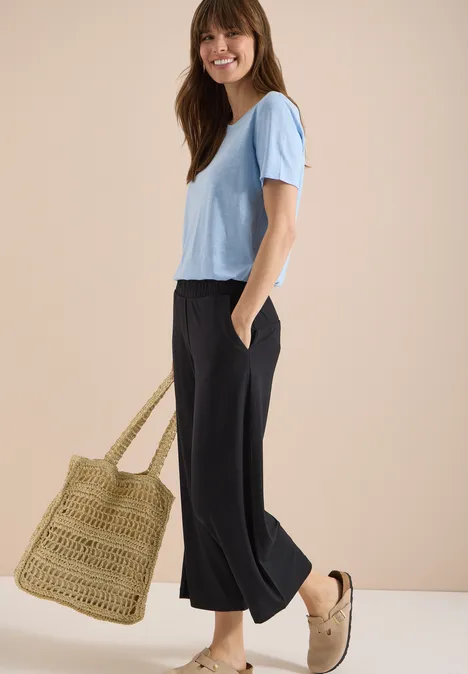3/4 Wide Leg Hose im Casual Fit Black Eine Person in einem hellblauen T-Shirt, schwarzen weiten Hosen, Basttasche und beigen Slippern.