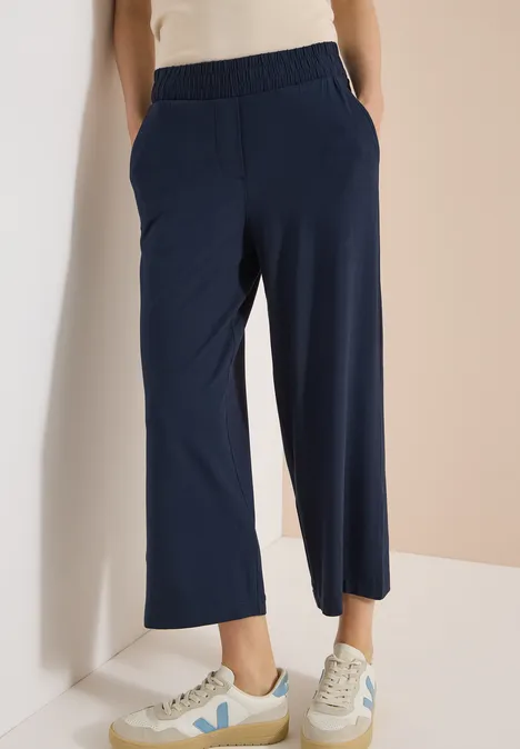 Dunkelblaue, weite Culotte-Hose mit weißen Sneakern vor hellem Hintergrund.