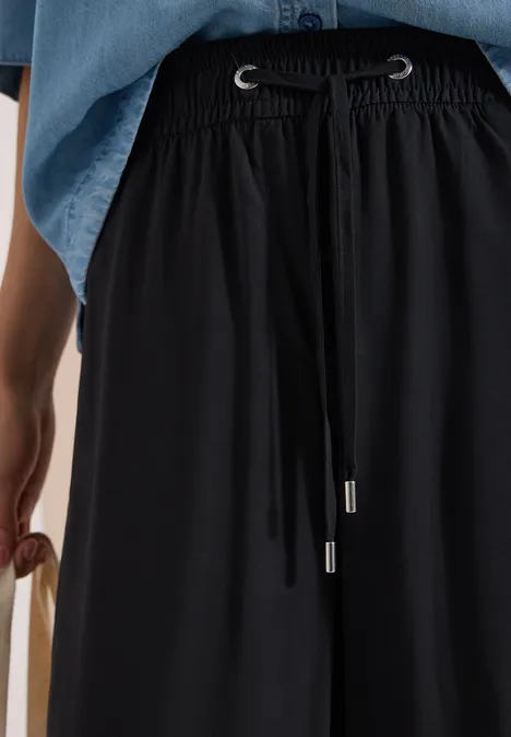 Schwarze, weite Stoffhose mit Tunnelzugbund, kombiniert mit Jeanshemd und seitlich gehaltener Tasche.
