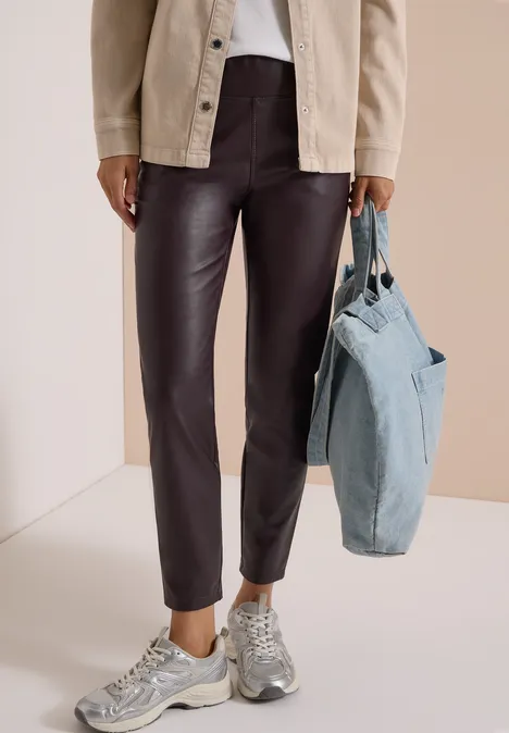 Eine Person trägt braune Lederhose, graue Sneaker, blaue Denim-Tasche und helle Jacke vor neutralem Hintergrund.