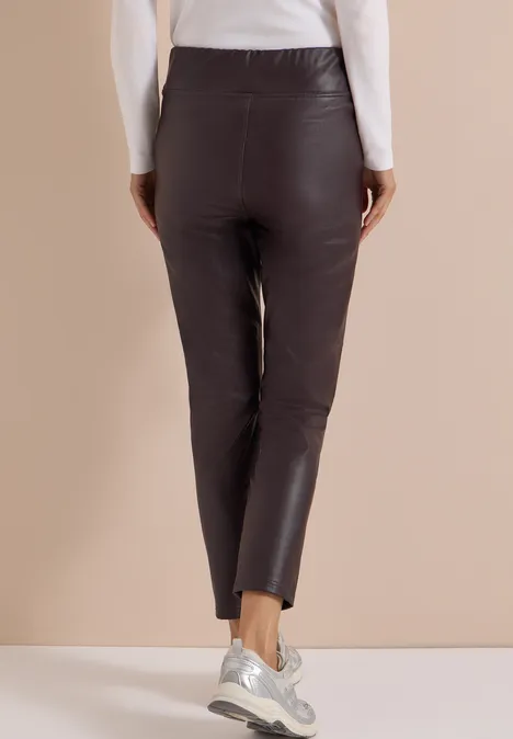 Rückenansicht: Person in schwarzen, enganliegenden Lederhosen und weißem Oberteil vor beigem Hintergrund.