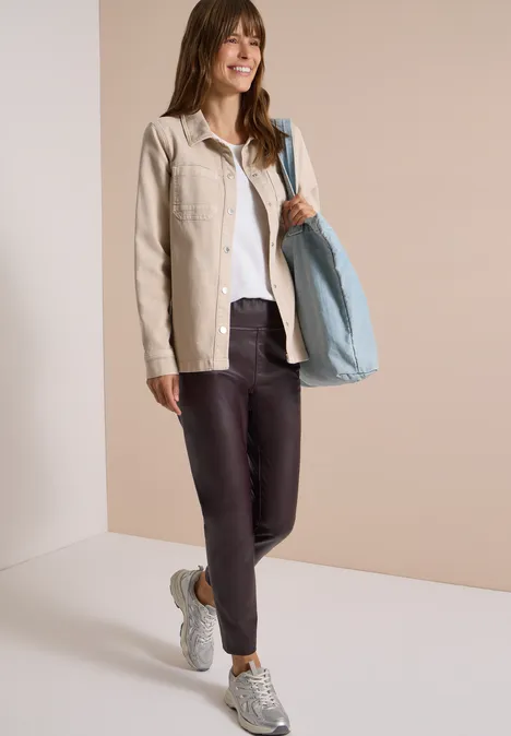 Frau in beiger Jacke, weißem Shirt, dunklen Leggings, Sneakern und großer hellblauer Tasche, die lächelnd posiert.
