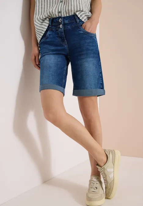 Person lehnt an Wand, trägt dunkelblaue Jeans-Bermuda und helle Sneaker