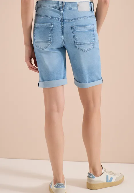 Persoon van achteren in lichtblauwe denimshorts met omslag en beige sneakers tegen neutrale achtergrond