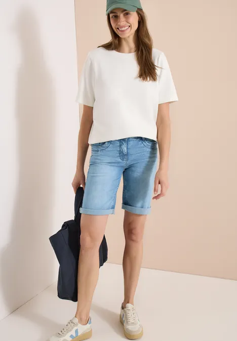 Vrouw draagt witte T-shirt, blauwe jeansbermuda en witte sneakers.