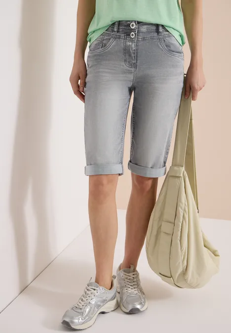 Femme en bermuda en jean gris clair retroussé, baskets argentées, top vert et grand sac en toile beige.