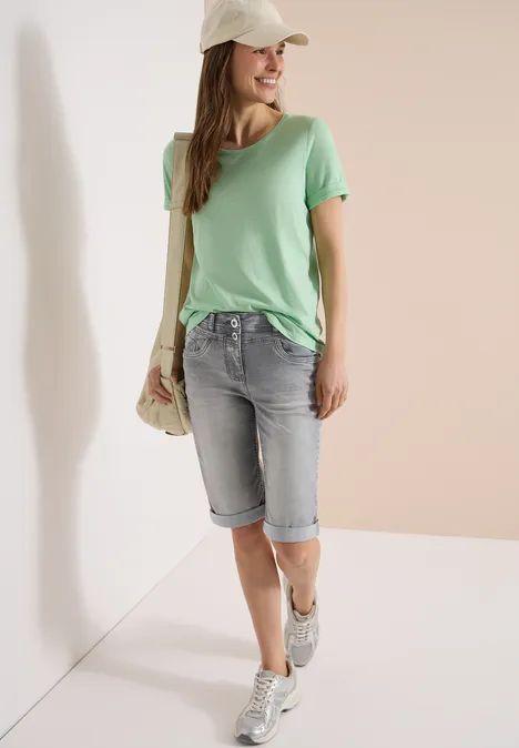 Une femme portant un t-shirt vert, un short en jean aux genoux, des baskets, un sac en paille et une casquette.