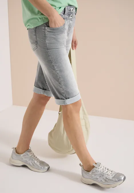 Jambes en bermudas bleu clair retroussés, baskets argentées, haut vert et sac en toile beige.