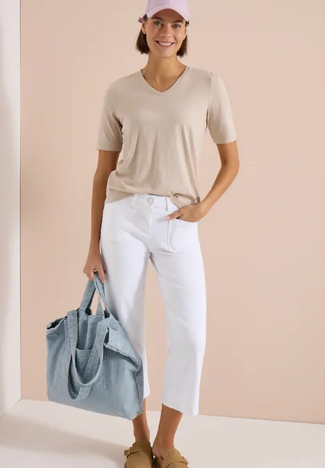 Vrouw in beige t-shirt, witte capri-jeans en grote lichtblauwe shopper voor een neutrale achtergrond.