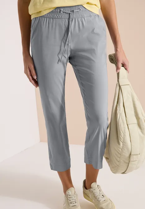 Vrouw in grijze 7/8-broek, lichte sneakers, met een gewatteerde tas in de hand.