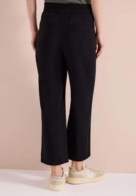 Frau von hinten in schwarzen, weiten, knöchellangen Hosen mit elastischem Bund und hellbeigen Sneakern.