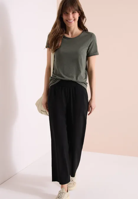 Frau in legerem Outfit: grünes T-Shirt, schwarze weite Hose und Sneaker vor hellem Hintergrund.