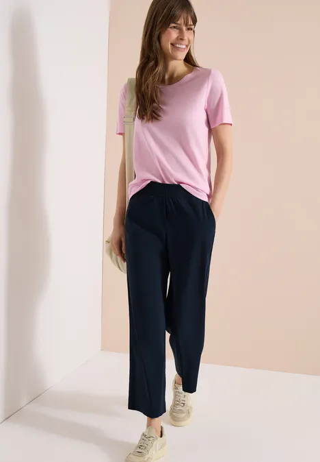 Vrouw in een loszittend roze T-shirt en donkerblauwe, hoog getailleerde broek voor een neutrale achtergrond.