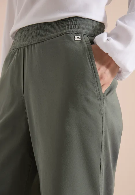 Olivgroene, losvallende broek met elastische taille, zijzak en klein logo, gecombineerd met wit shirt.