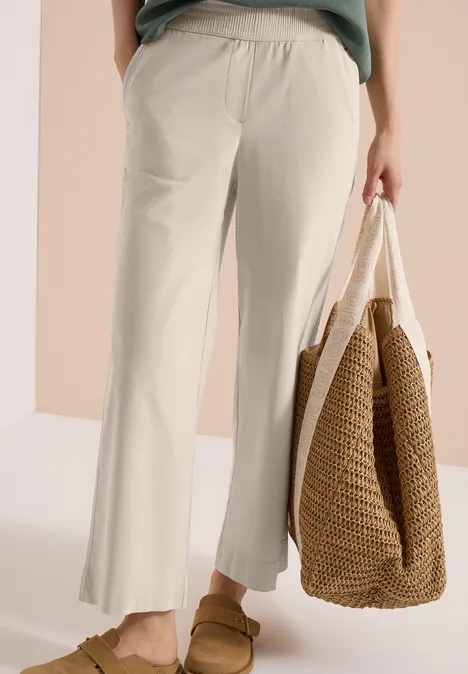 Beine in beigen, weiten 7/8-Hosen mit Rüschensaum, braunen Pantoletten und großer geflochtener Tragetasche.