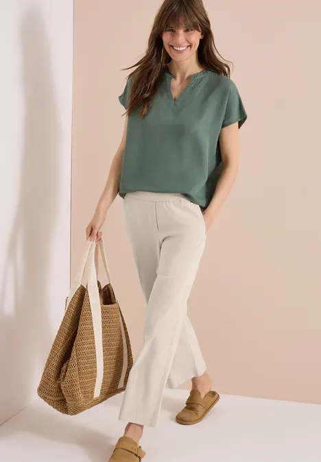 Frau trägt grünes Shirt, helle Schlaghose, Basttasche und Sandalen, steht vor hellem Hintergrund.