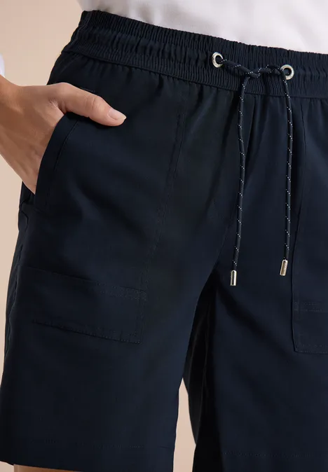 Dunkelblaue, lässige Hose mit Kordelzug und aufgesetzten Taschen, Hand in der Tasche.