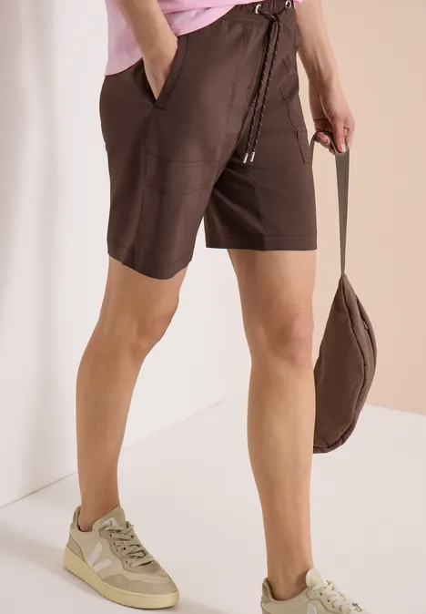 Person trägt braune Shorts, rosa Oberteil, beige Sneaker und hält eine passende braune Tasche.