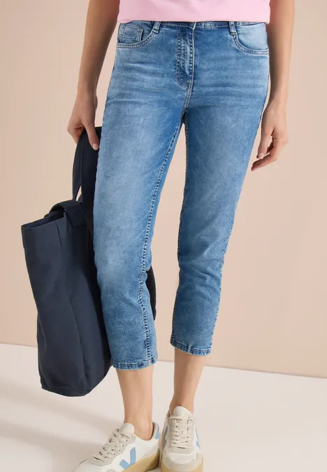 Vrouw draagt blauwe cropped jeans en witte sneakers, met donkerblauwe tas voor een beige achtergrond.