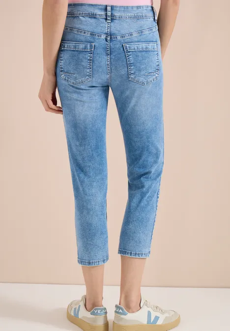 Vrouw van achteren in lichtblauwe, hoge taille jeans tot net boven de enkel, met witte sneakers voor een beige muur.