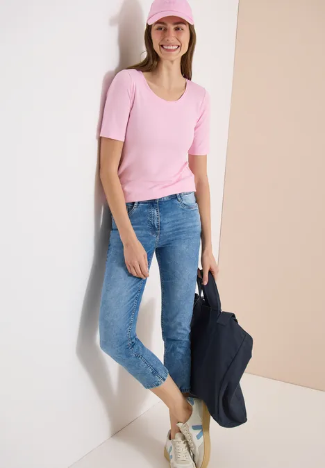 Iemand leunt tegen een witte muur, draagt een roze T-shirt, een blauwe jeans en houdt een donkere jas vast.