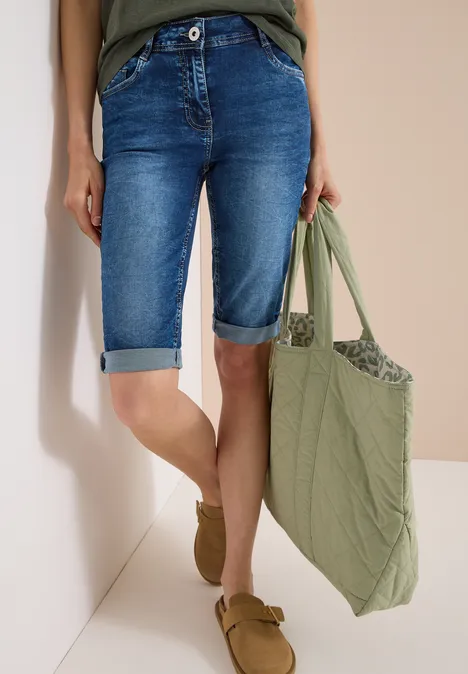 Person trägt blaue Denim-Bermudashorts, olivgrüne Tasche und braune Slipper vor neutralem Hintergrund.