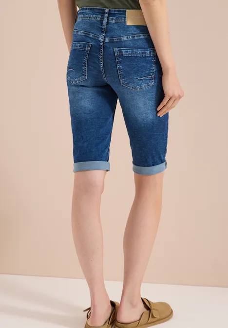 Modell trägt blaue, umgeschlagene Denim-Bermuda-Shorts und olivegrüne Slipper vor neutralem Hintergrund.