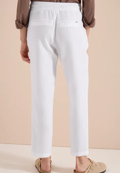 Rückansicht einer Person in weißen Leinenhosen und beigen Loafers vor hellem Hintergrund.