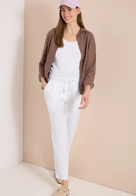 Frau in weißer, lässiger Jogginghose und Top mit kastiger, brauner Jacke vor neutralem Hintergrund.