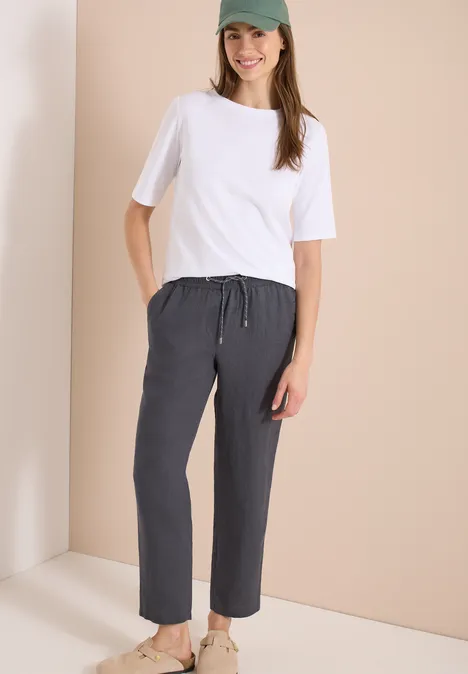 Vrouw in wit t-shirt en losse donkergrijze broek voor een beige achtergrond.