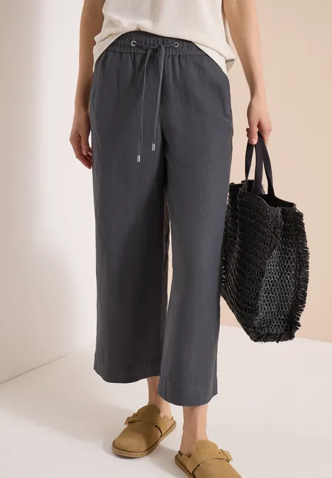 Person mit grauer, weiter Leinenhose, weißem Top, goldenen Hausschuhen und schwarzer Flecht-Tasche.