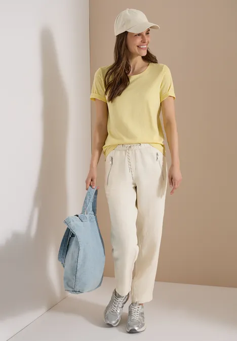 Femme en t-shirt jaune, pantalon beige, casquette et baskets grises, portant un sac bleu.