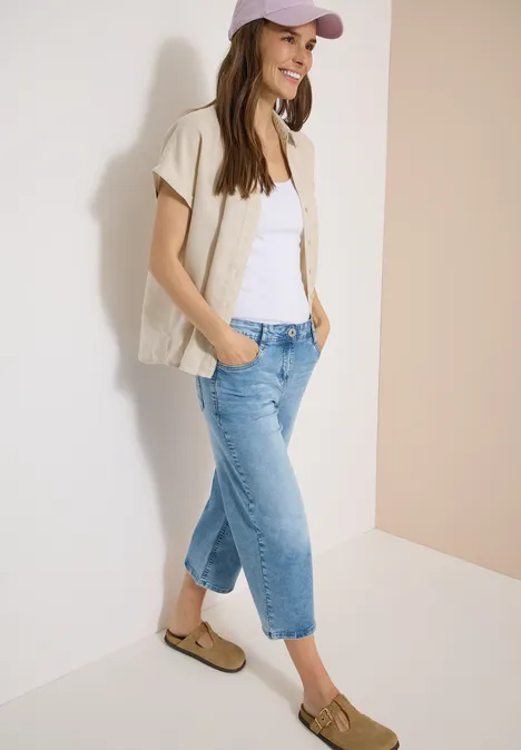 3/4 High Waist Wide Leg Jeans im Slim Fit light blue washed Junge Frau lehnt lässig an Wand, trägt beiges Kurzarmhemd, weißes Top, blaue Jeans, Loafer und Cap.