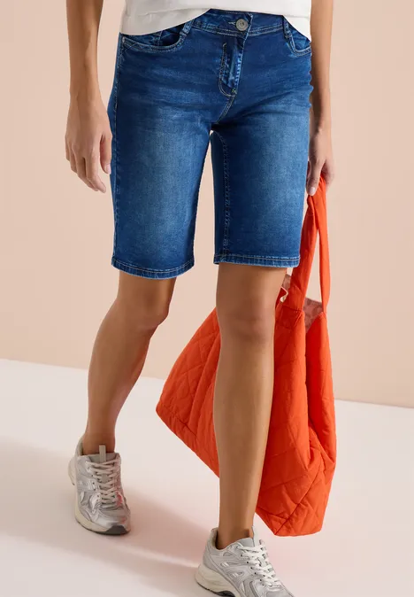 Vrouw draagt knielange spijkershorts, witte sneakers en houdt een oranje gewatteerde jas vast.